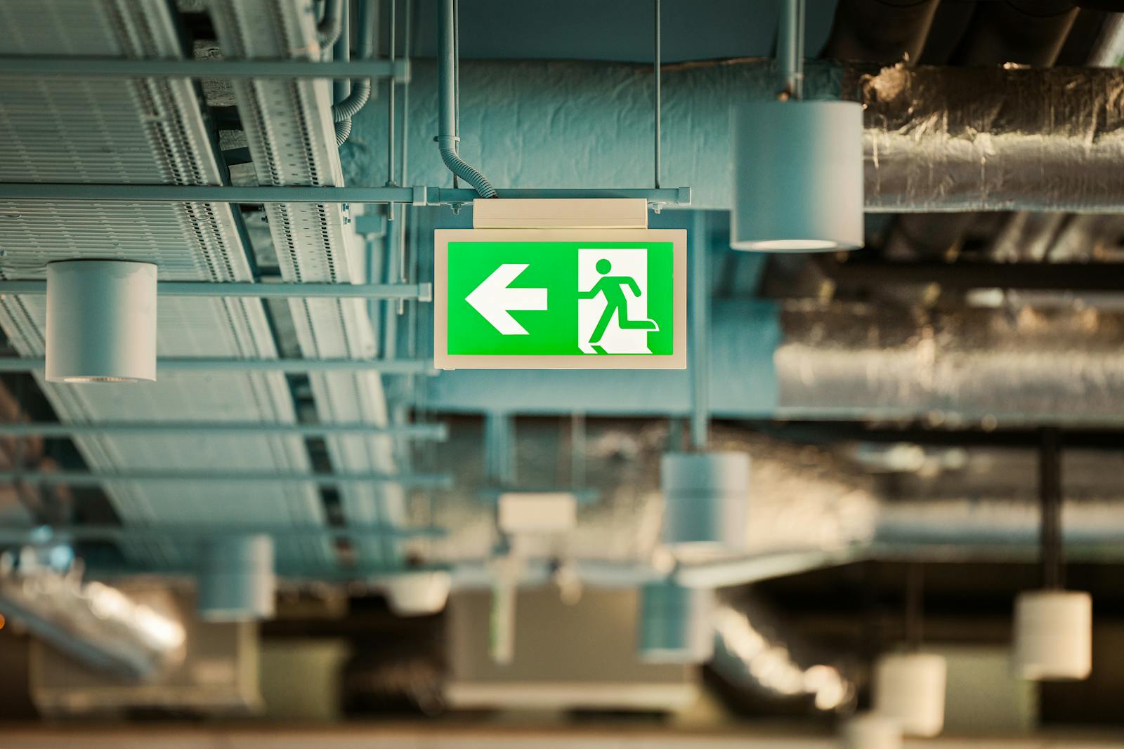 Stairwell pressurization testing — green emergency exit sign in a commercial corridor with exposed ductwork above, pressurized egress under NFPA 92